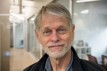 Portrait of a smiling older man with gray hair and beard in a modern office hallway.