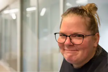 Smiling woman with glasses in an office setting, glass walls visible in the background.